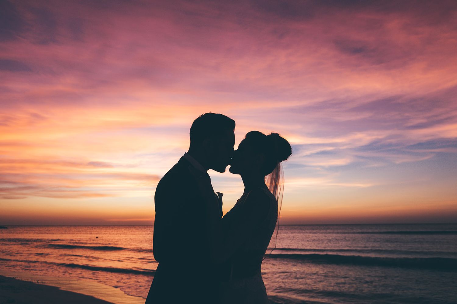 Silhouettes of a couple against a vibrant pink and orange sunset sky over the ocean.