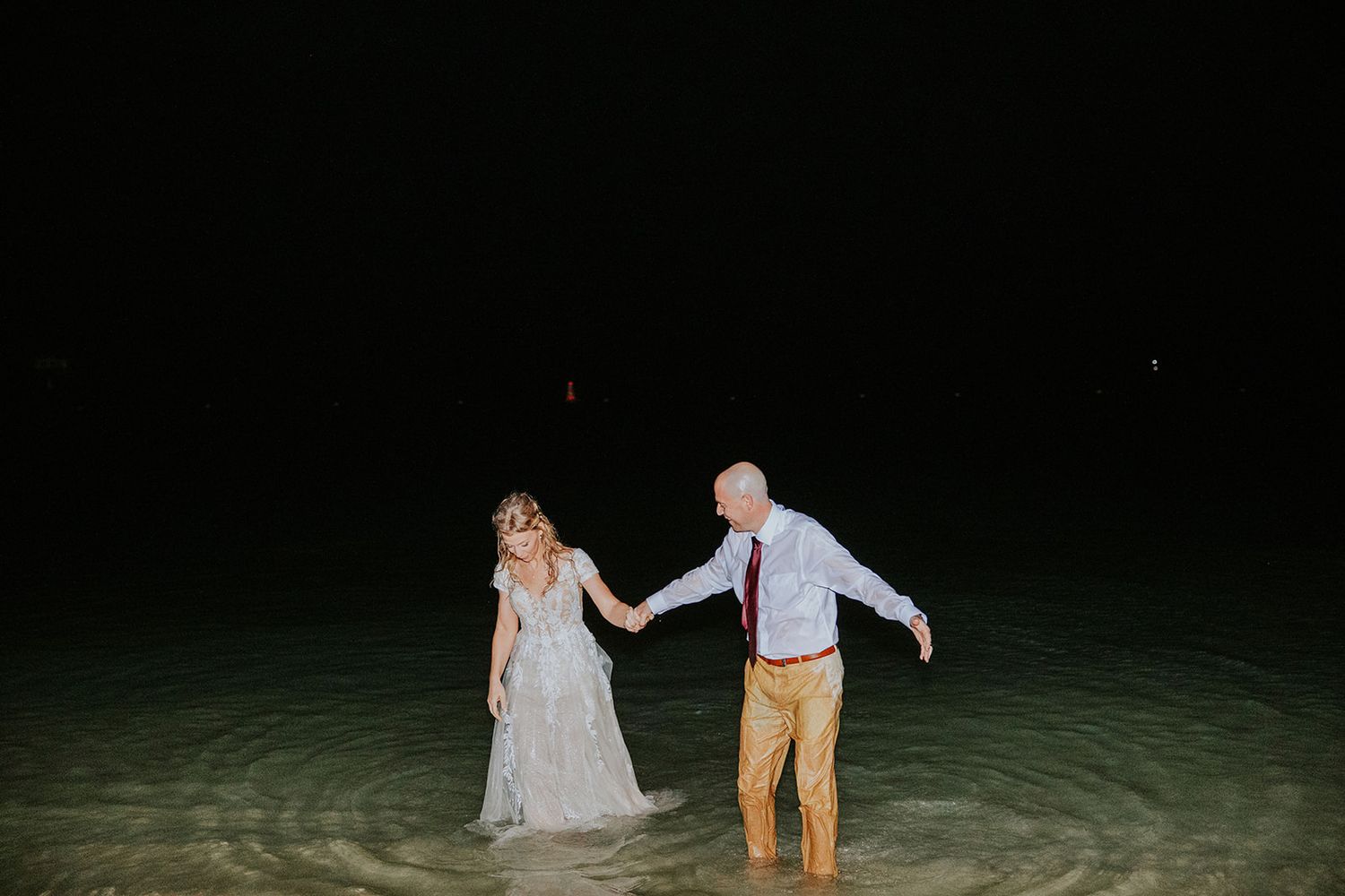 Newlyweds walk hand in hand along a moonlit beach at night in their wedding attire.