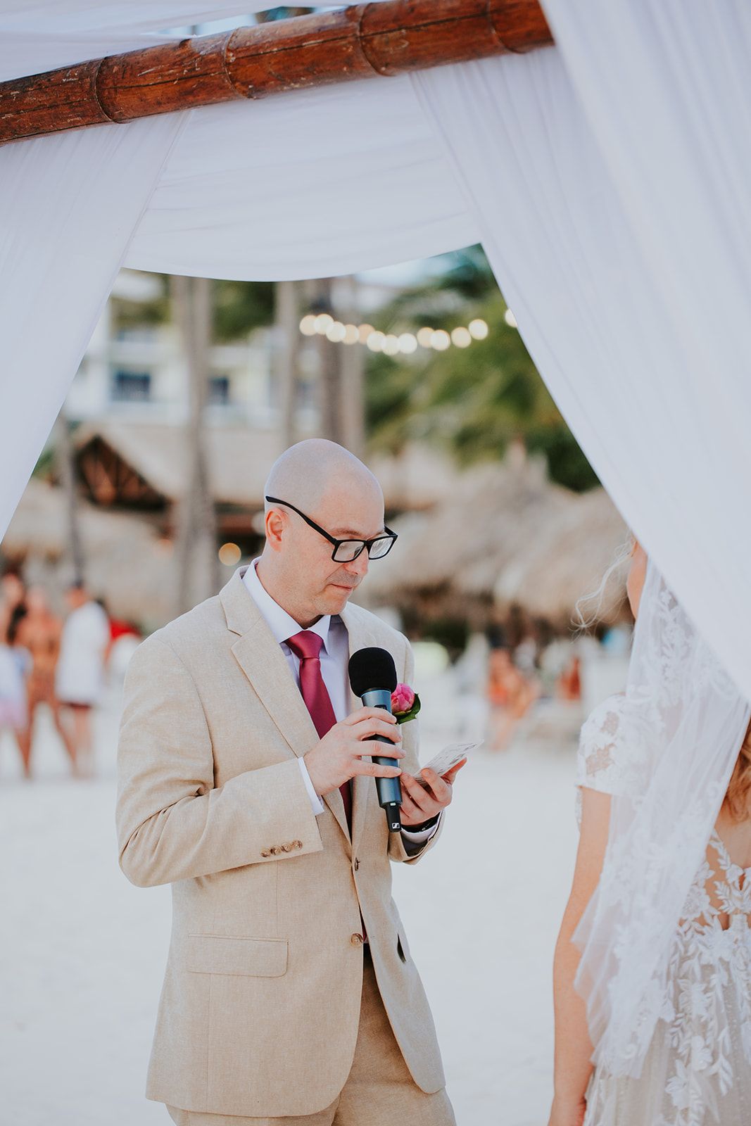 Wedding officiant in beige suit and red tie reads from phone during beach ceremony under wooden arch.