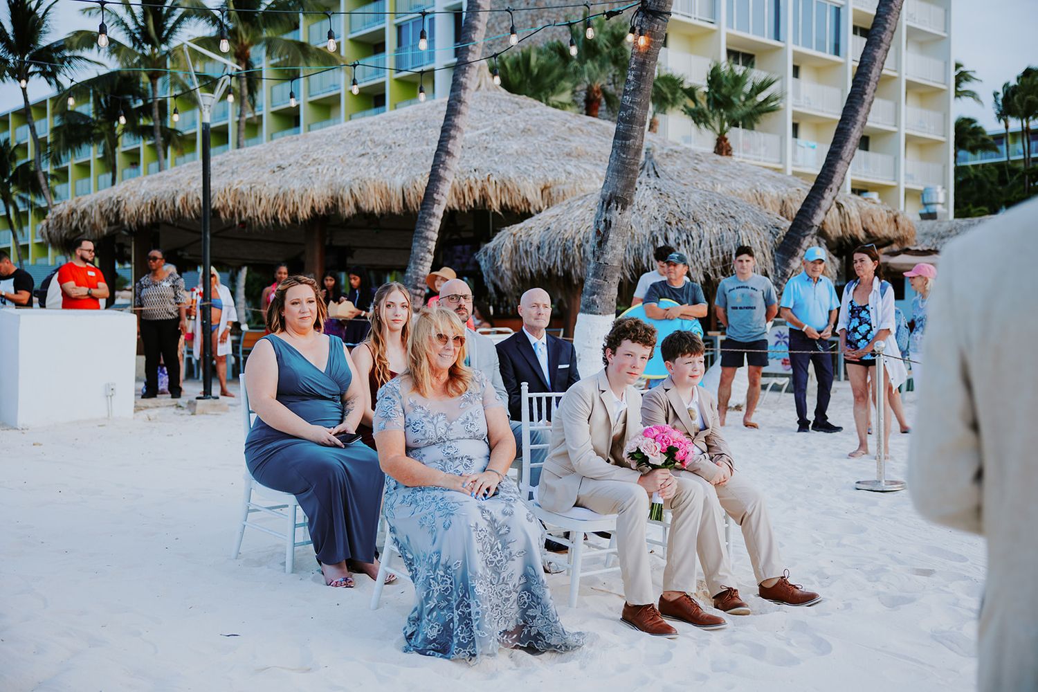 Wedding guests in casual attire gather on a sandy beach during a tropical destination ceremony.