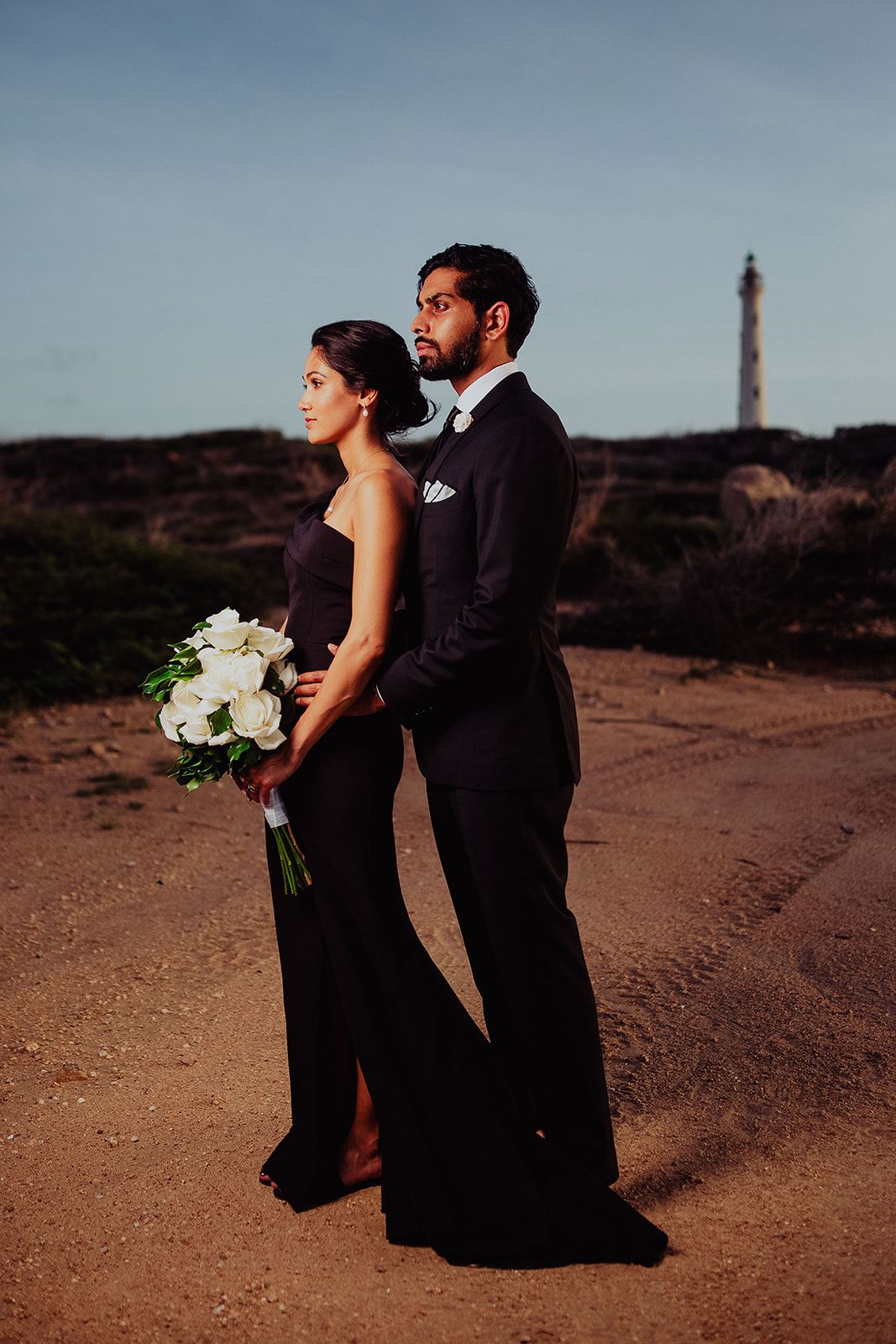 A formal couple poses at sunset with a lighthouse in the background.