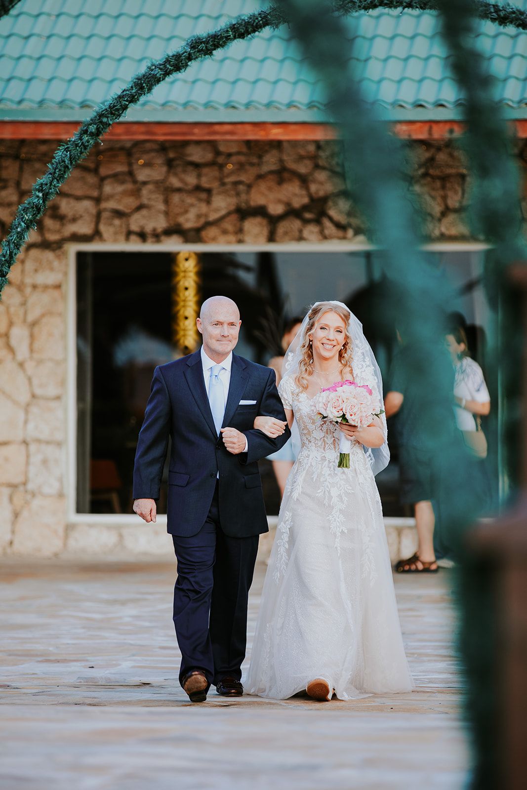 A couple in formal wedding attire walks down a stone pathway under green arches at an outdoor venue.