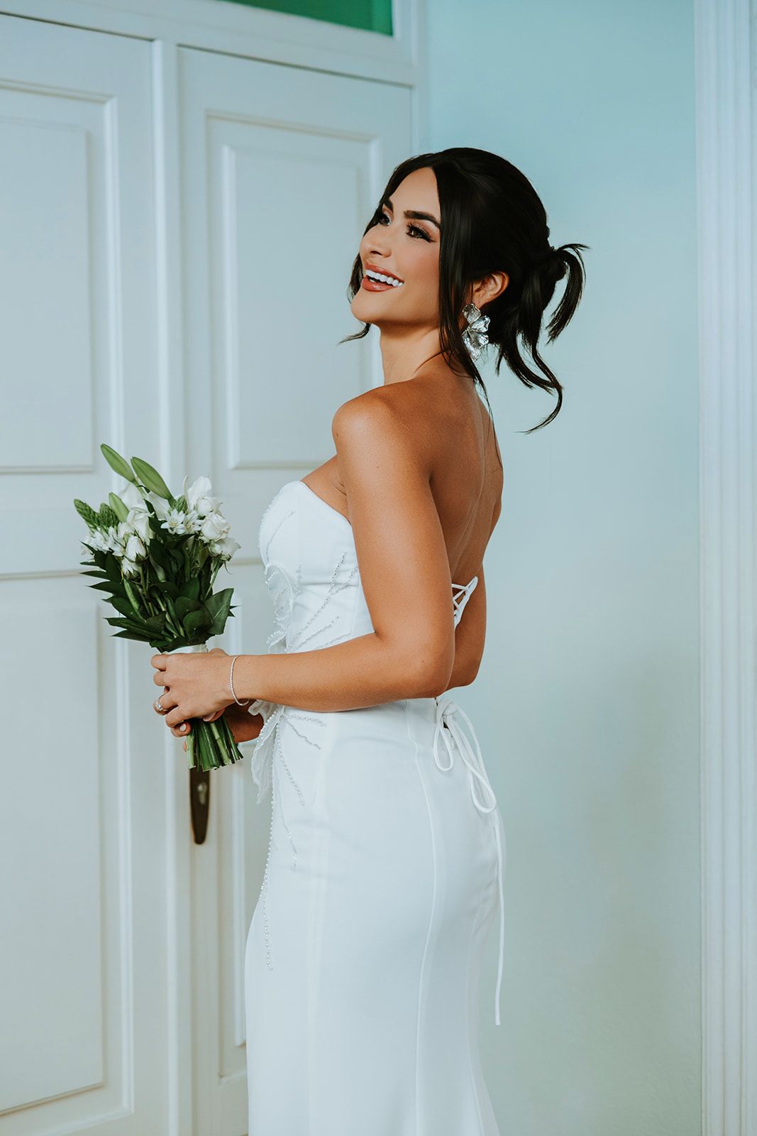 A bride in a white strapless gown poses elegantly in a bright room with white walls and floral accents.