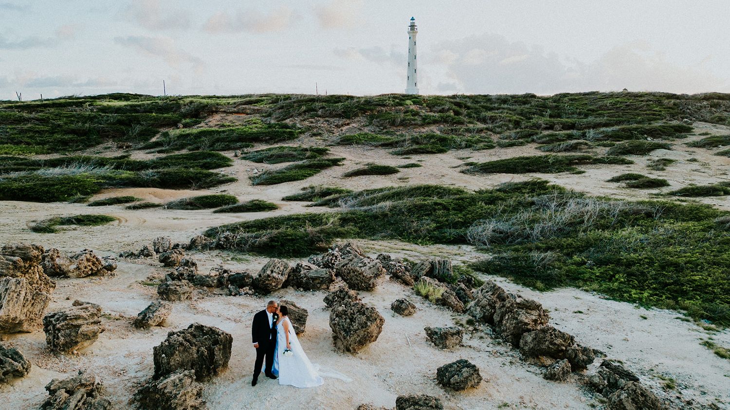 Wedding couple standing on rocky coastal terrain with lighthouse in background.