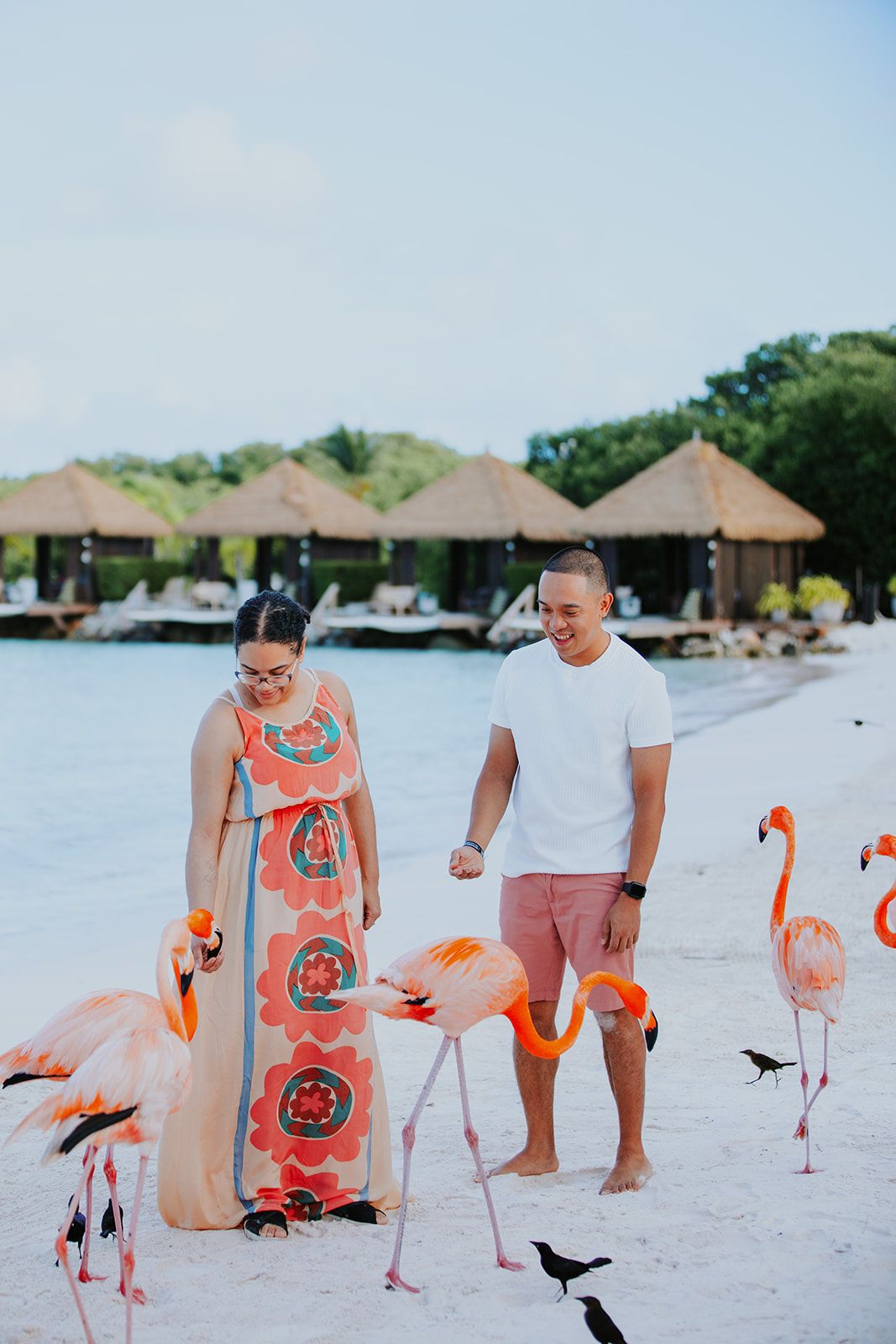Couple walking on white sandy beach with pink flamingos in the foreground at a tropical resort destination.