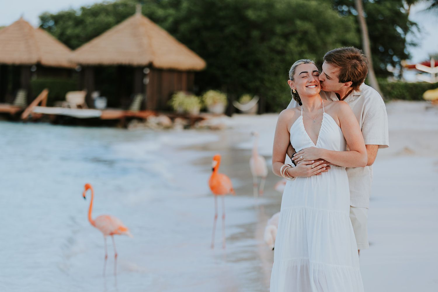 Group celebrating on a beach with palm trees and thatched huts at golden hour.