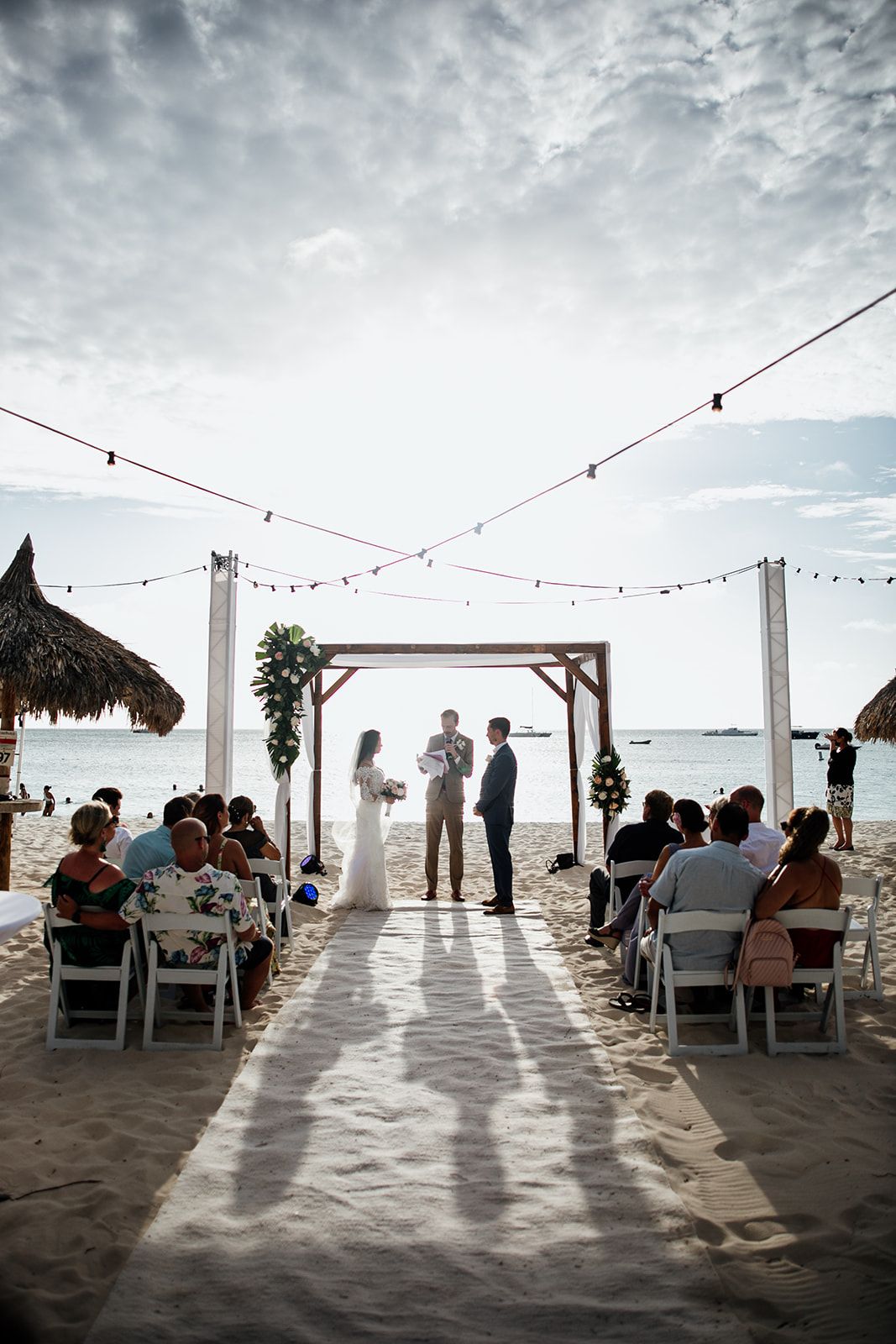 A beach wedding ceremony with guests seated in white chairs under a wooden arch by the ocean.