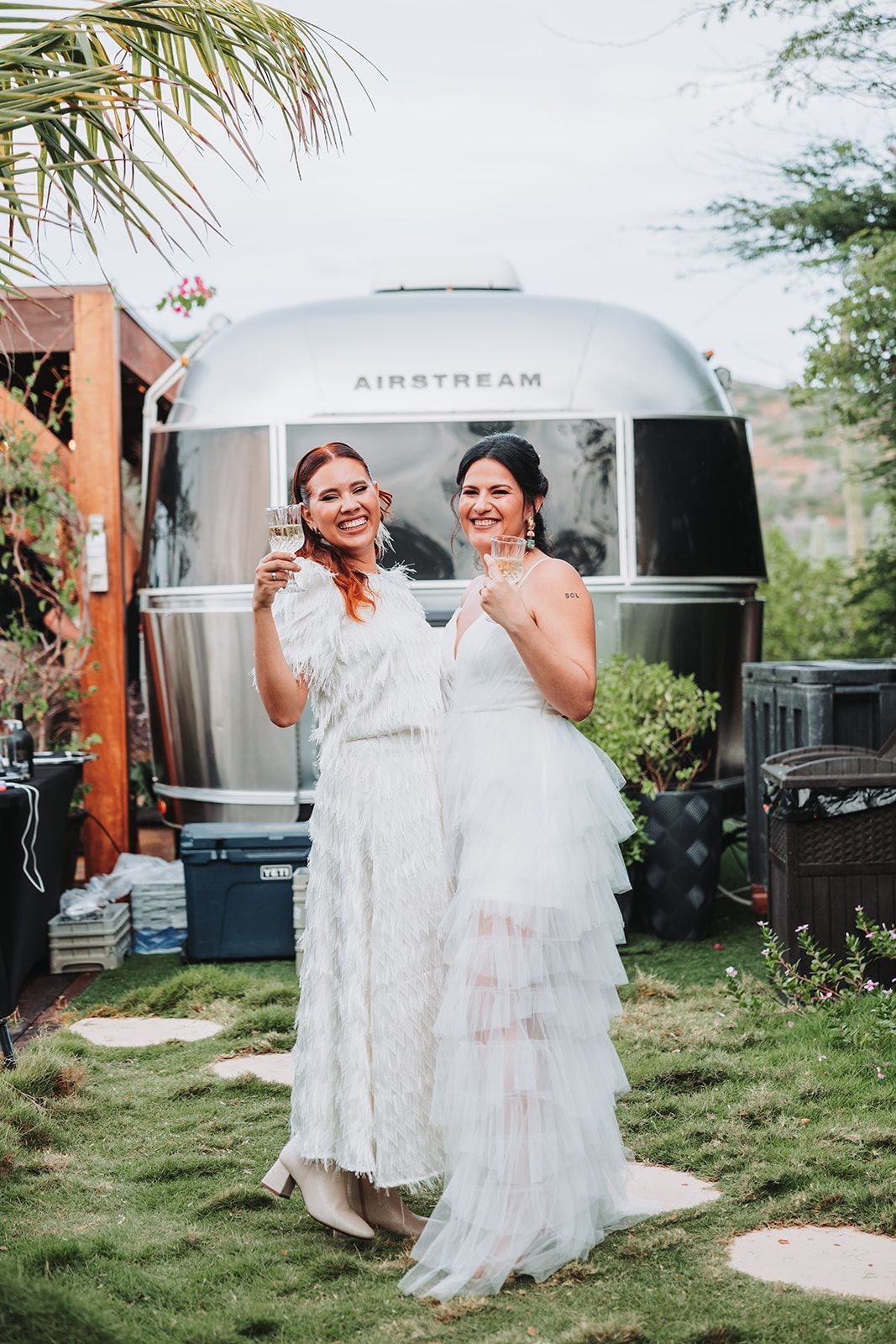 Two people in white wedding dresses laugh and dance together in front of a silver Airstream trailer on a grassy lawn.