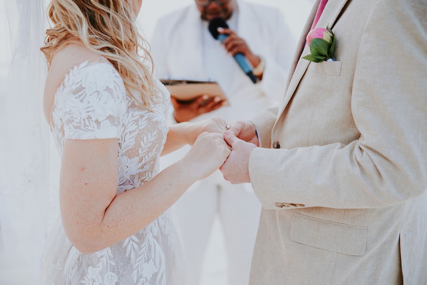 Elegant wedding couple holds hands during outdoor ceremony with sunlight filtering through white floral decorations.