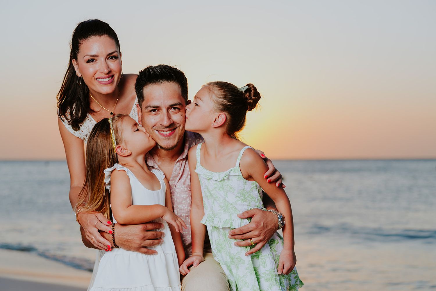 Family of four smiling at sunset on a beach, two daughters kissing their father on the cheeks.