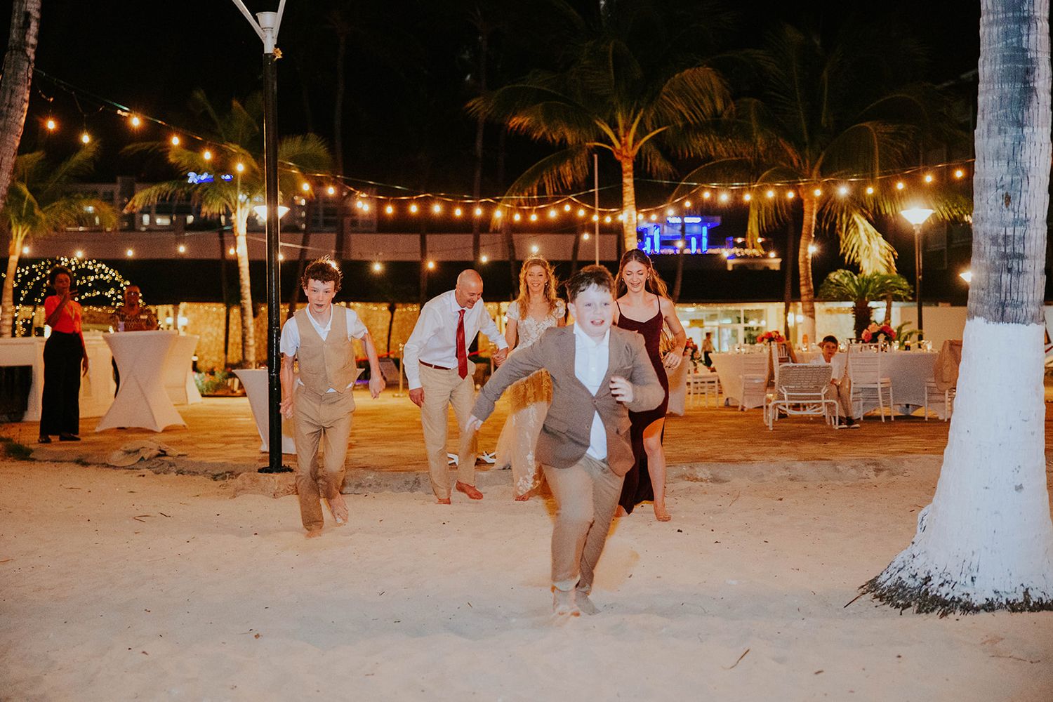 Wedding guests dance and celebrate on a sandy beach at night under warm string lights and palm trees.