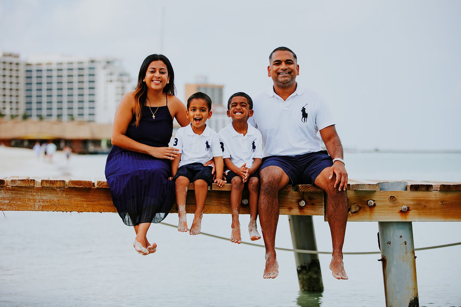 A family sits together on a wooden pier at the beach with a hotel visible in the background during Aruba sunset