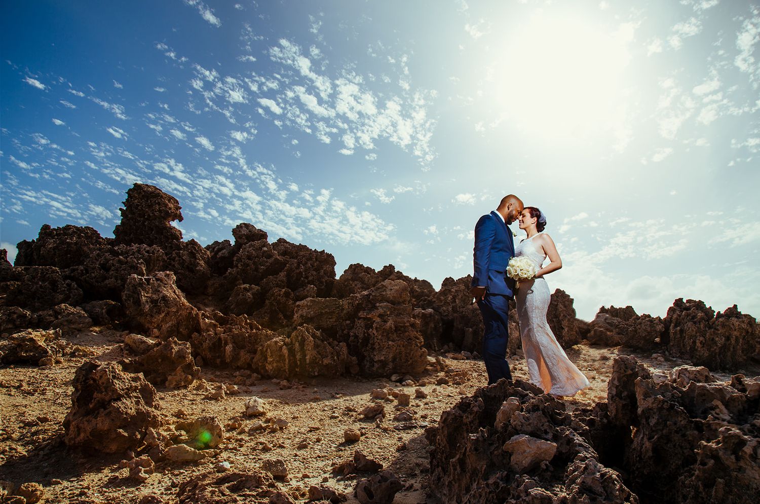 A couple stands atop dramatic coastal rock formations under a bright blue sky with wispy clouds.