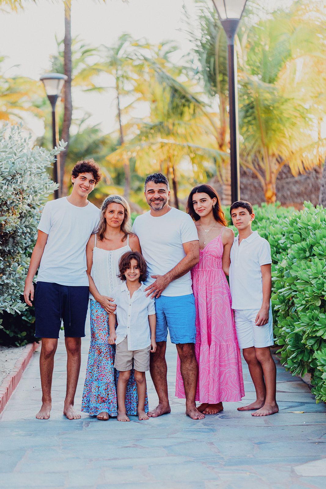 Happy family in casual beach attire poses together on a palm-lined pathway.