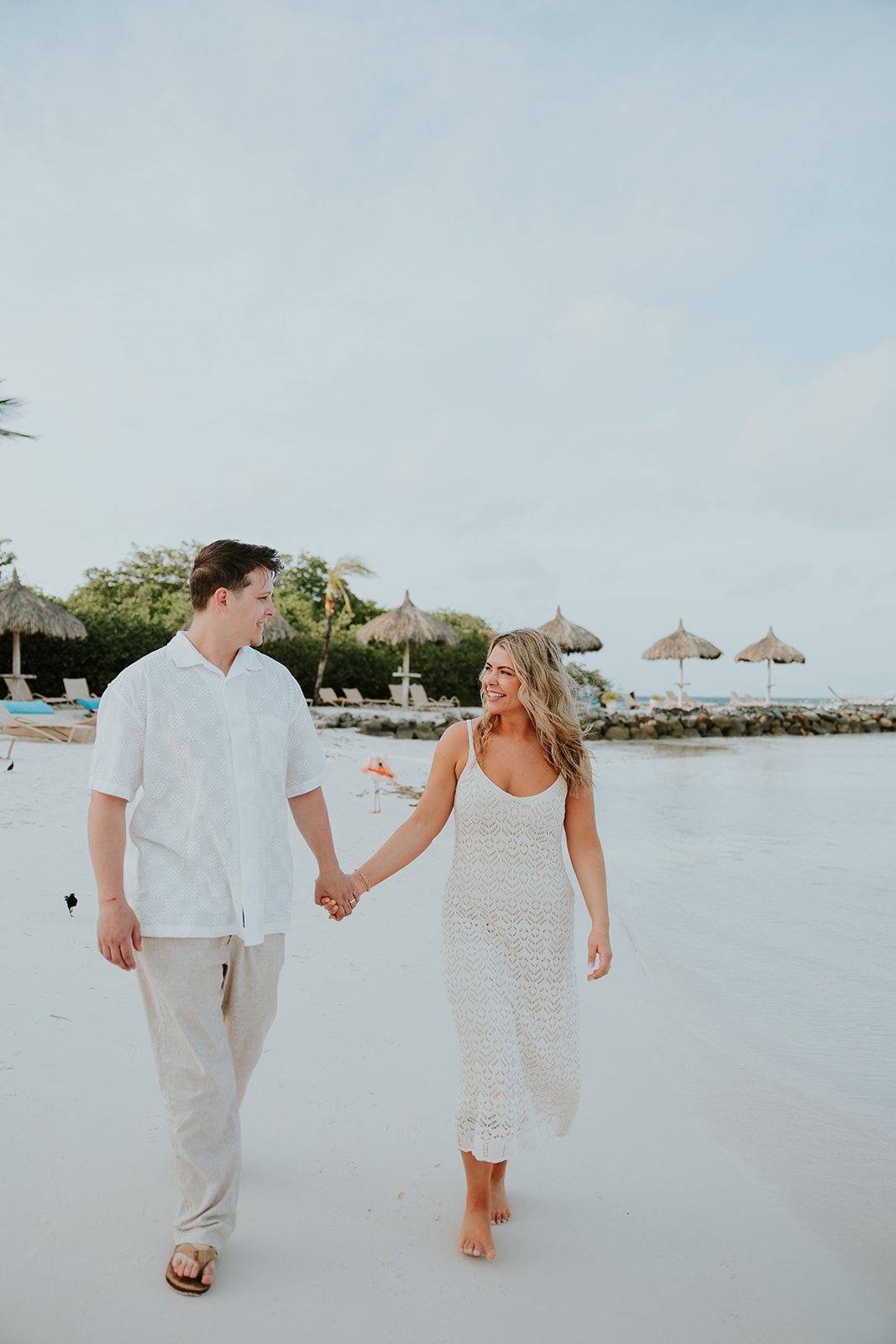 A couple walks hand in hand along a tropical beach with thatched umbrellas and palm trees in the background.