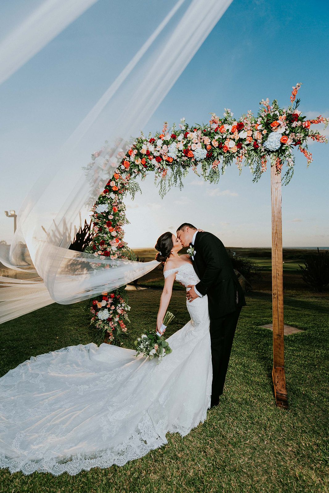 Wedding ceremony setup with floral arch and long flowing veil caught in breeze.