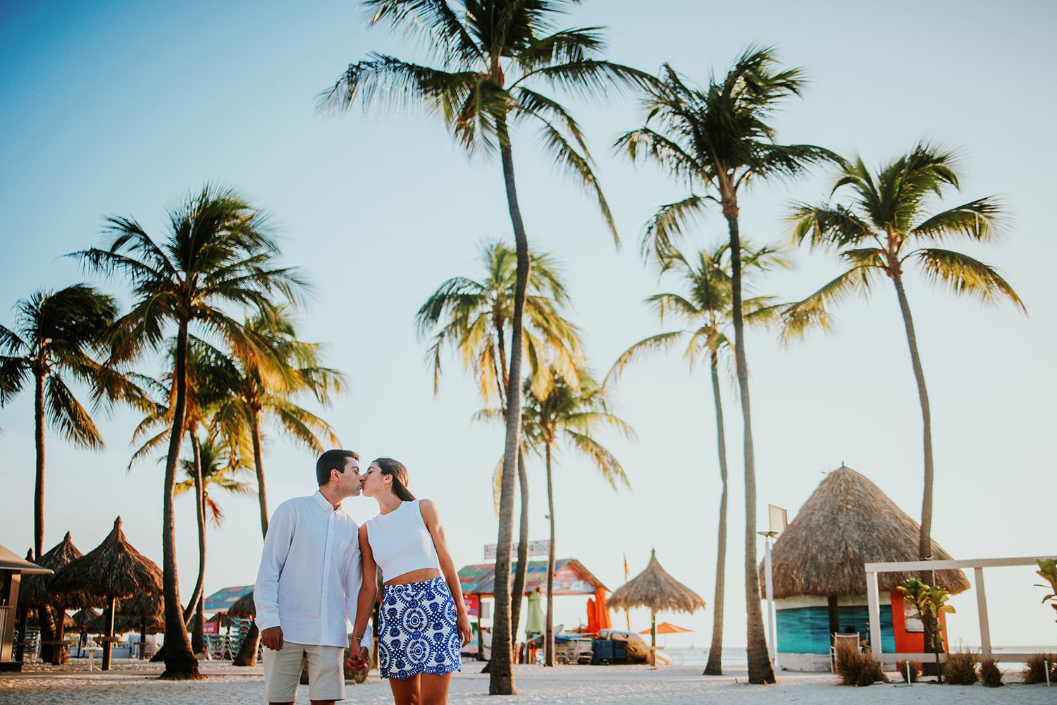Tropical beach wedding ceremony setup with palm trees and thatched umbrellas at sunset.