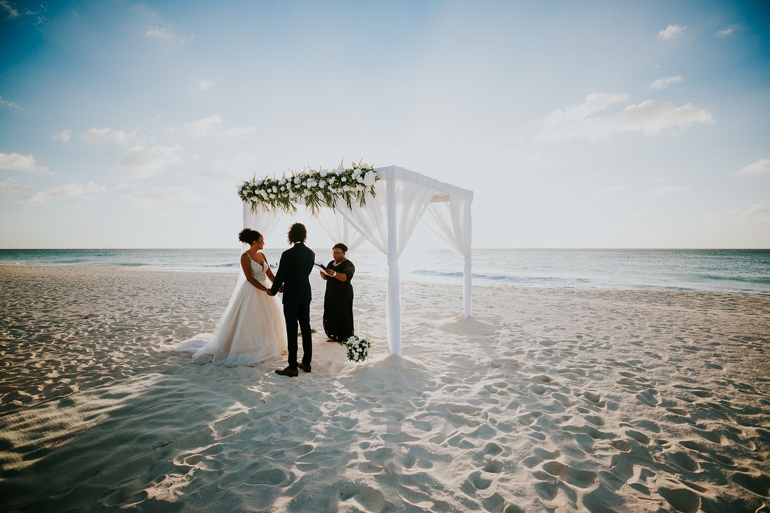 Aruba beach wedding ceremony under a white arch decorated with flowers at sunset.