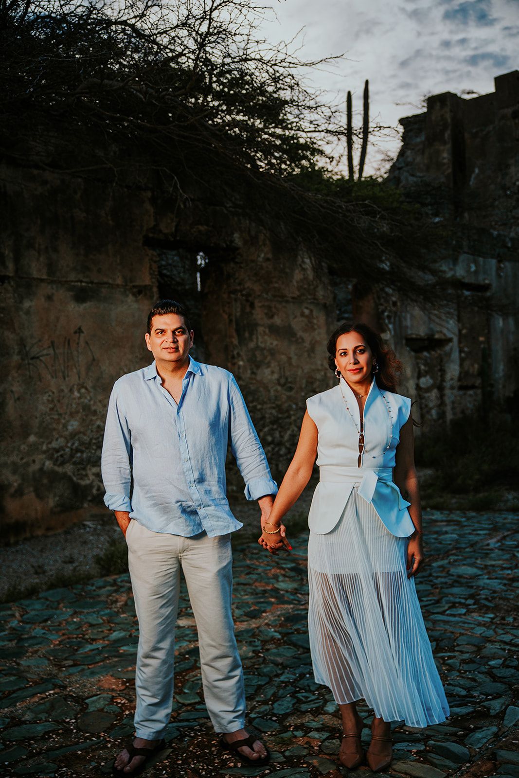 A couple in light blue and white outfits poses together in an outdoor setting at sunset.