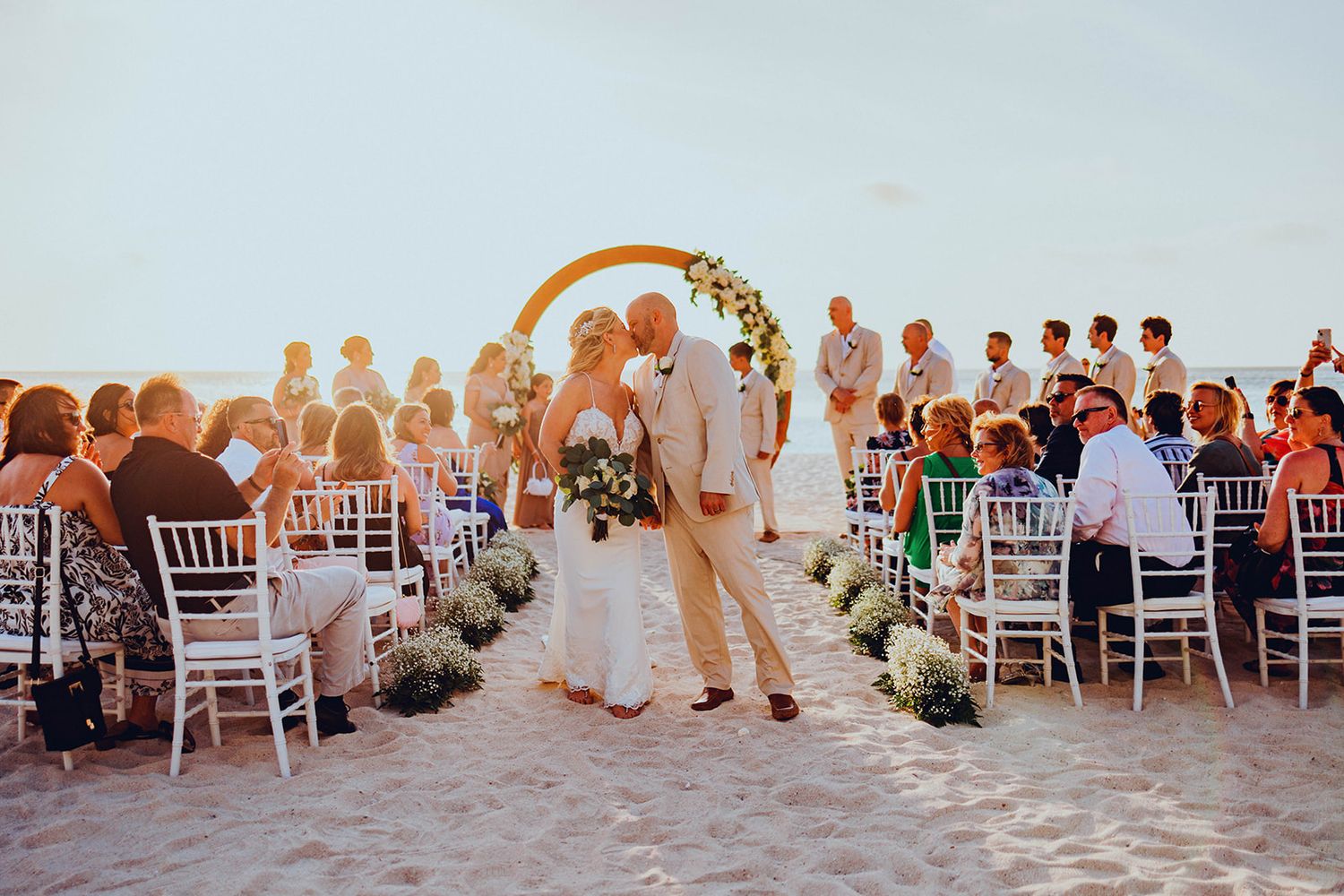 Wedding guests celebrate on a sunny beach with an elegant arch and white chairs during a romantic sunset ceremony.