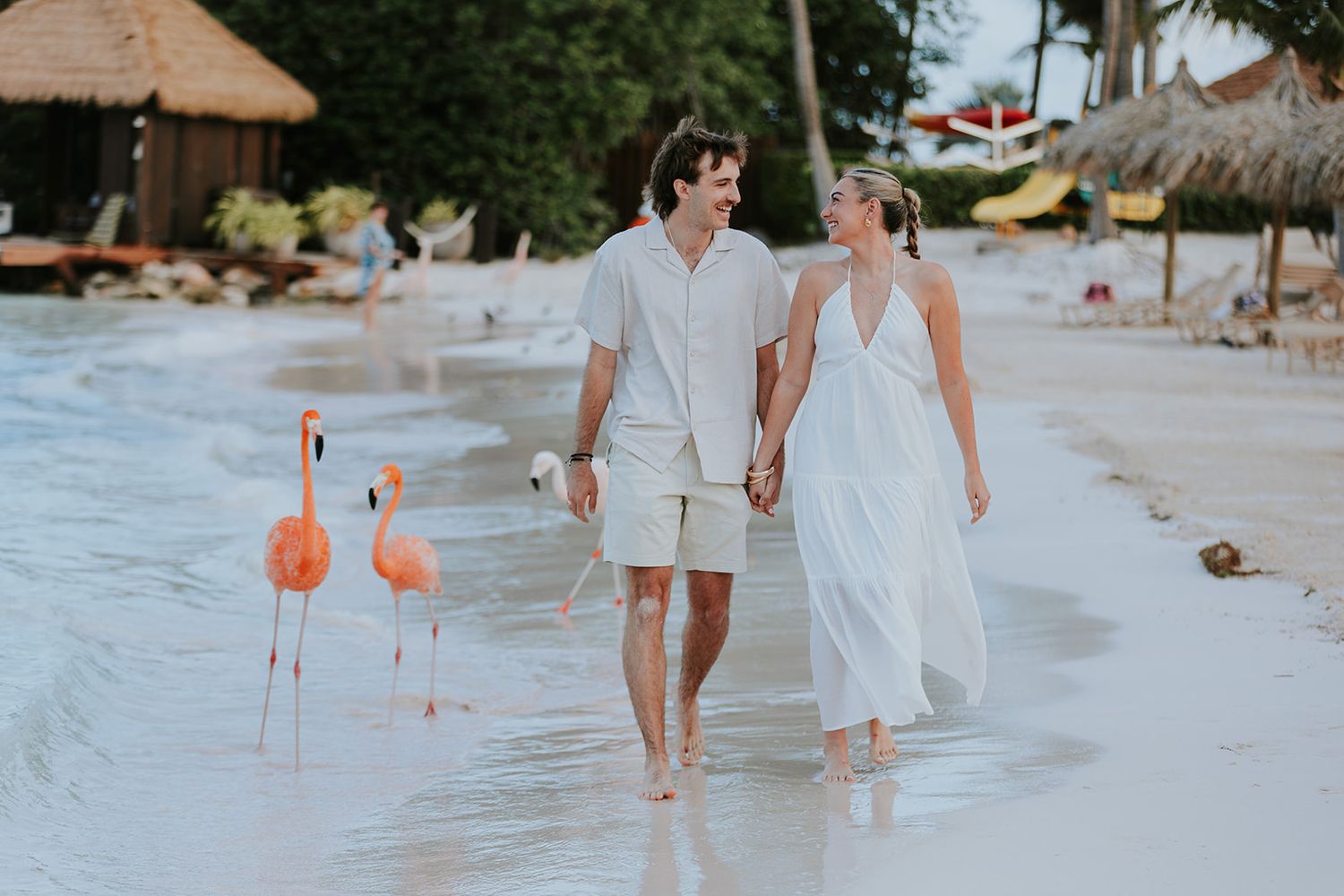 Couple walking along a tropical beach with thatched umbrellas and pink flamingo decorations in the sand.