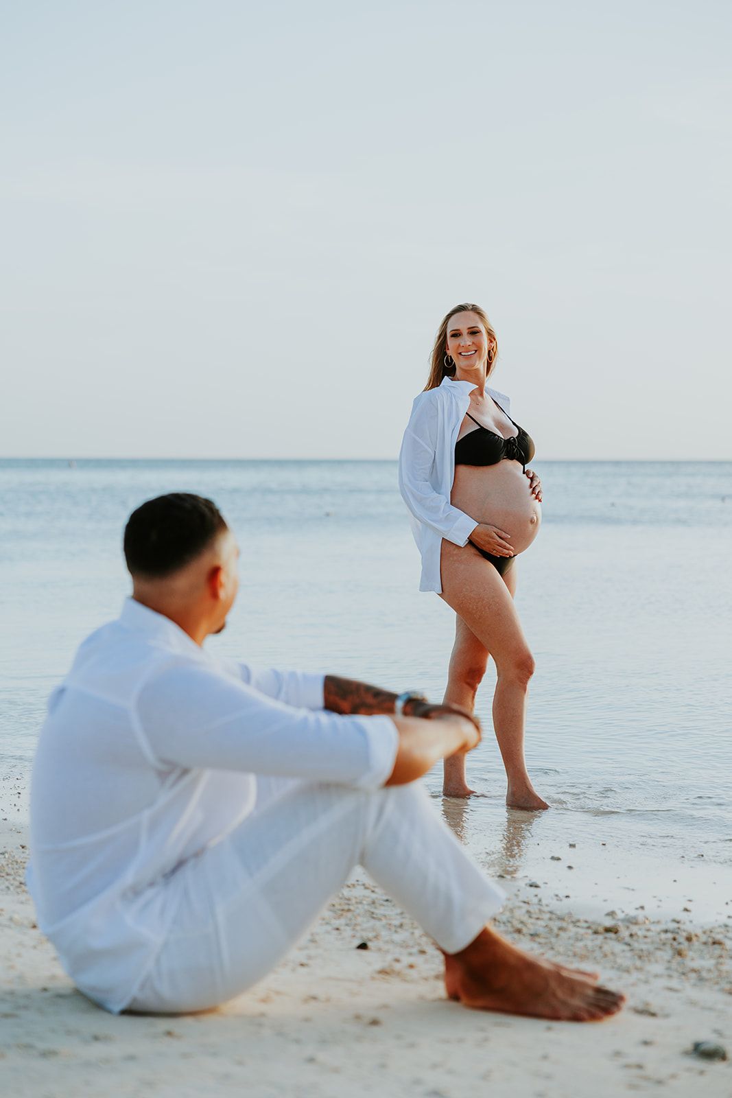 Maternity beach photoshoot with couple sitting in white attire by the ocean.