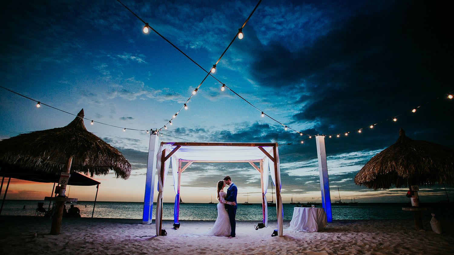 A beachfront wedding ceremony under string lights and tiki huts at twilight.