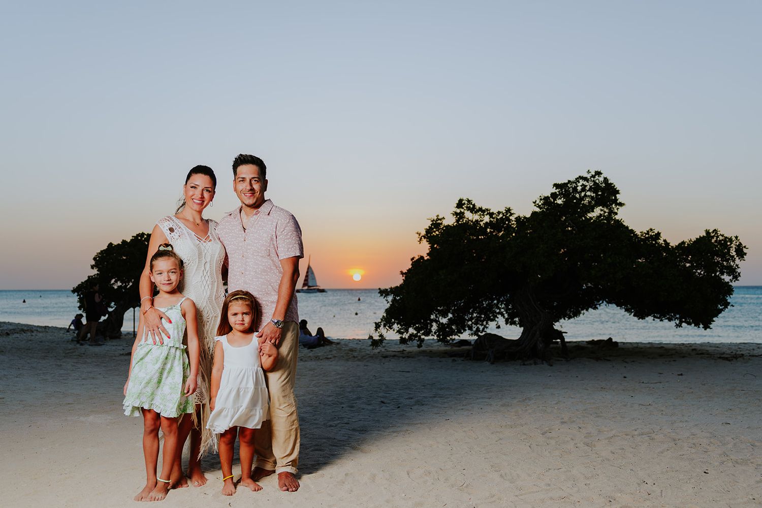 People pose together on a tropical beach at sunset with palm trees and ocean in the background.