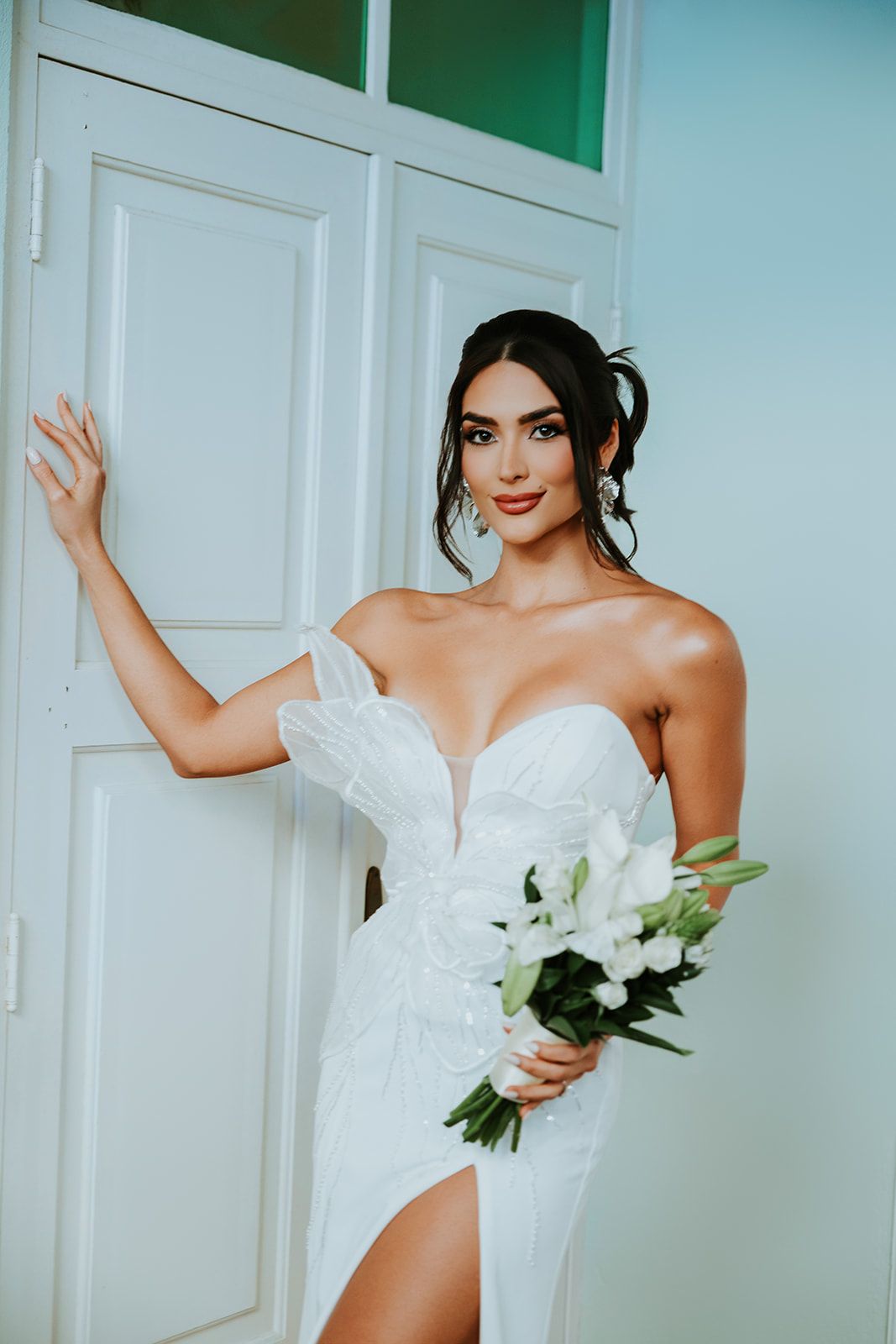 A bride in a strapless white wedding dress holds white lilies while posing against a mint green wall.