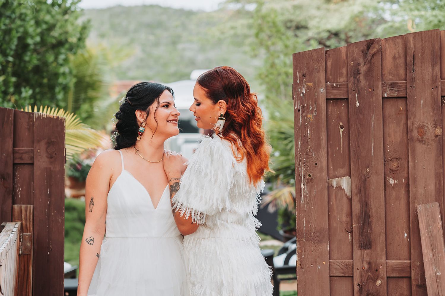 Two people in wedding attire embrace in front of a rustic wooden wall at an outdoor venue.