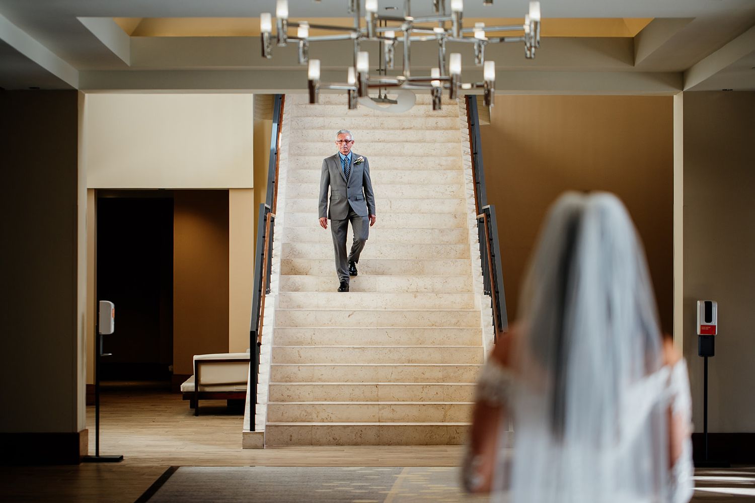 A wedding first look moment captured on an elegant indoor staircase under a modern chandelier.