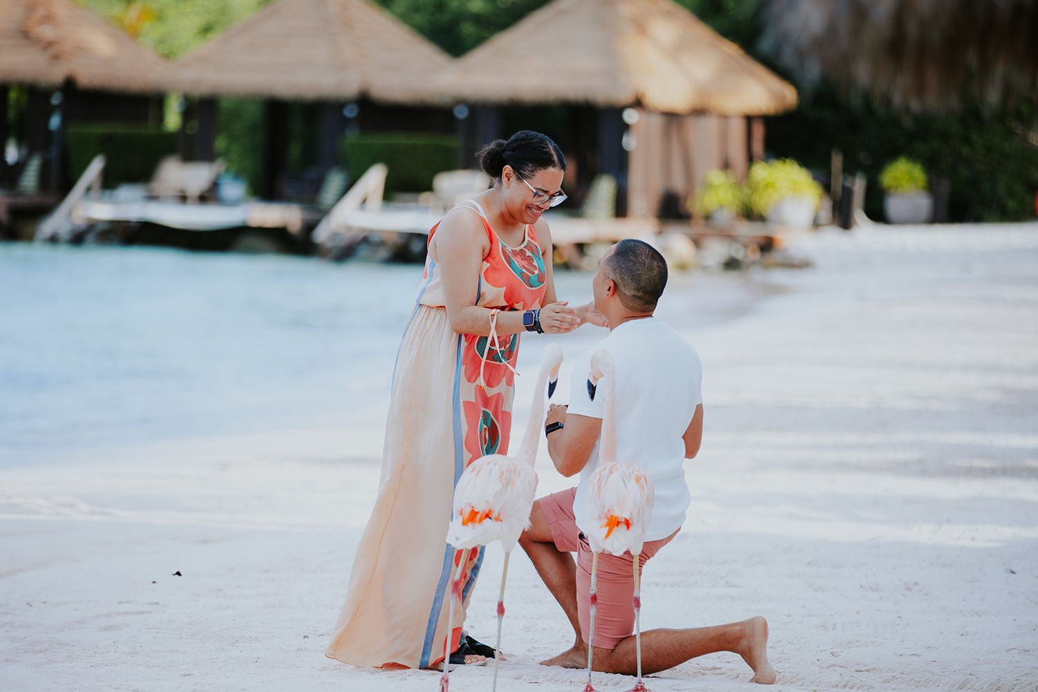 A romantic beach proposal scene with a couple in white and floral attire on a tropical resort beach at sunset.