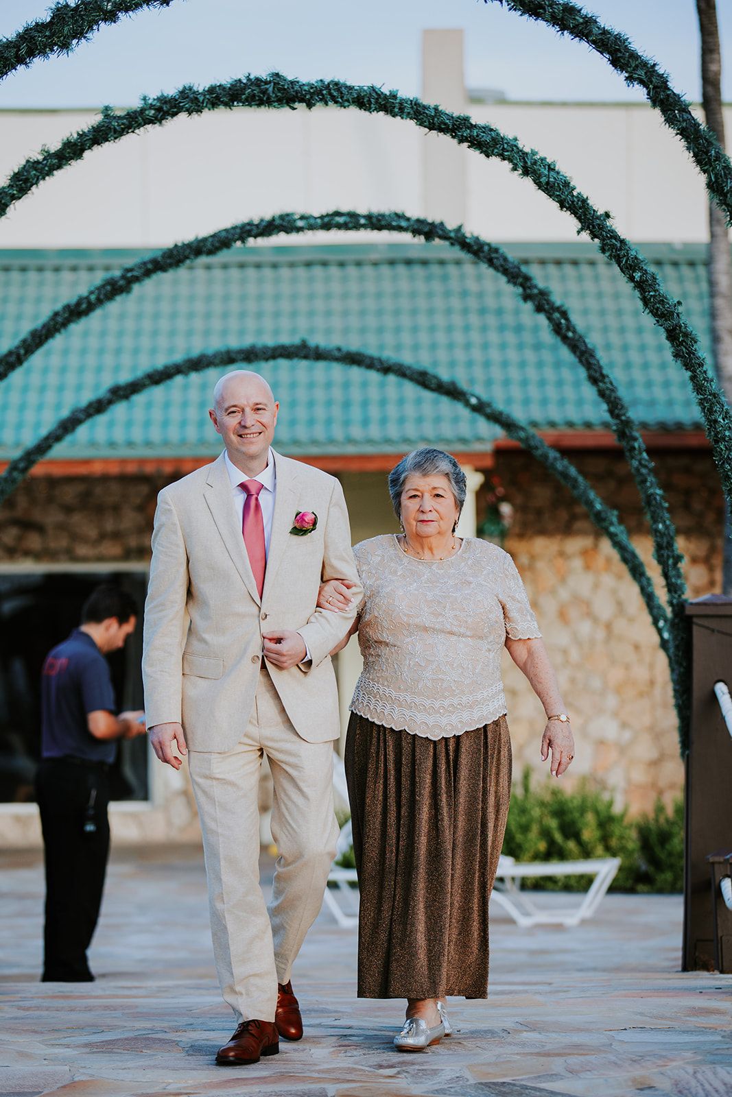An elderly couple walks together under decorative green arches in a courtyard with stone walls and teal roofing.