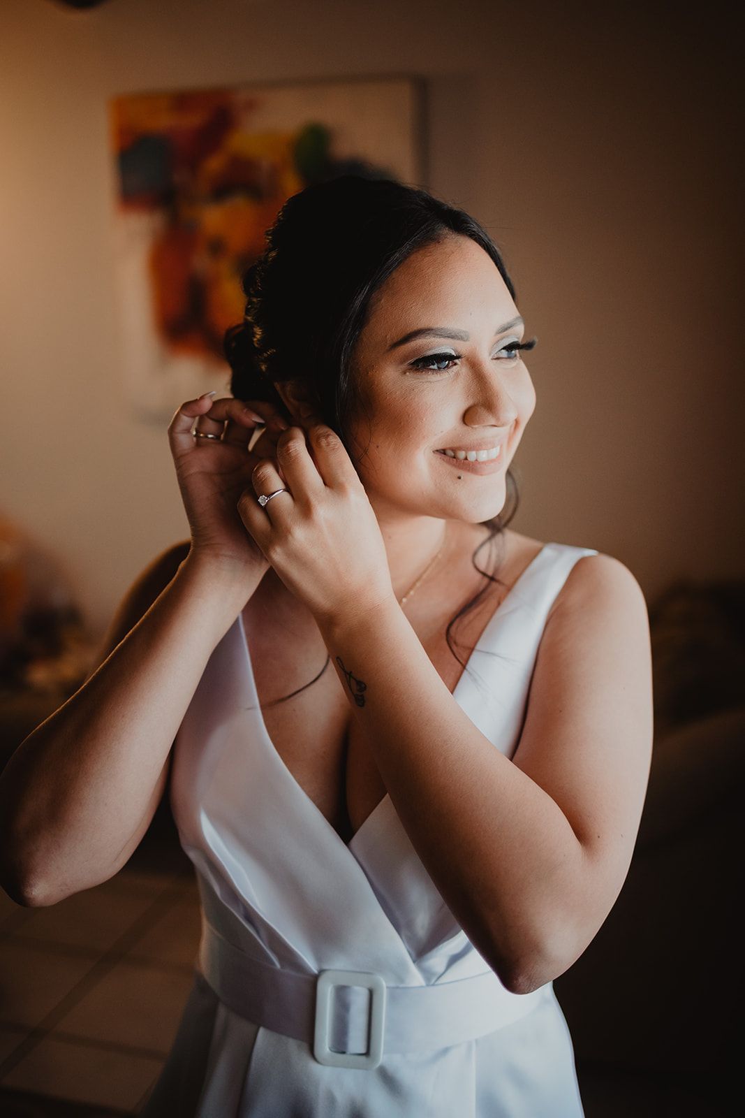 Person in white dress putting on earrings in warm indoor lighting.