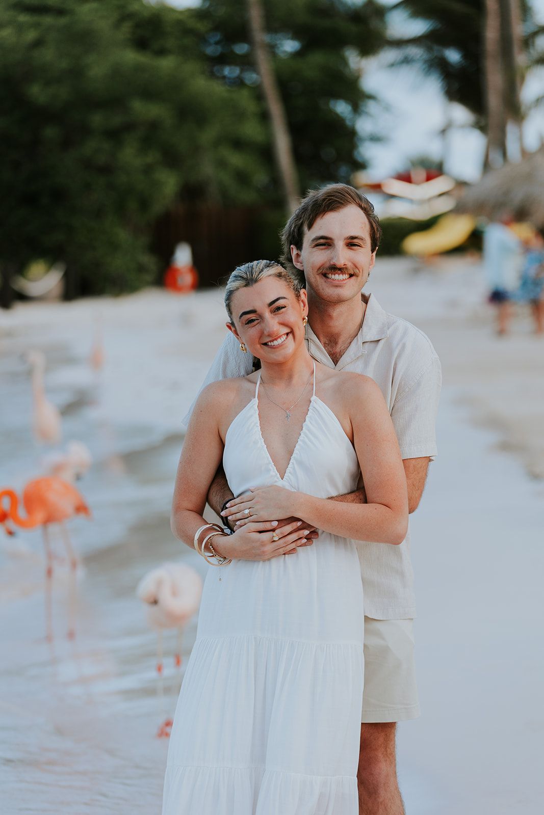 A couple embraces on a sunny beach, wearing white and beige outfits while sharing a romantic moment.
