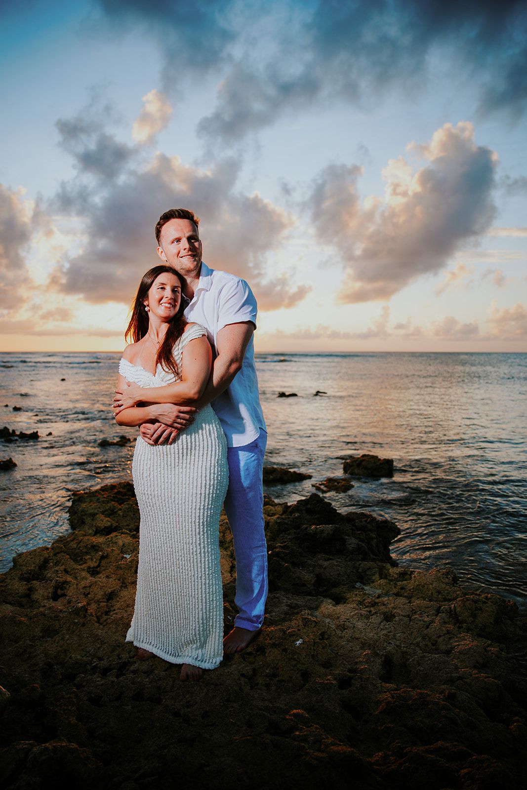 Couple embraces on rocky beach shore during golden hour sunset.