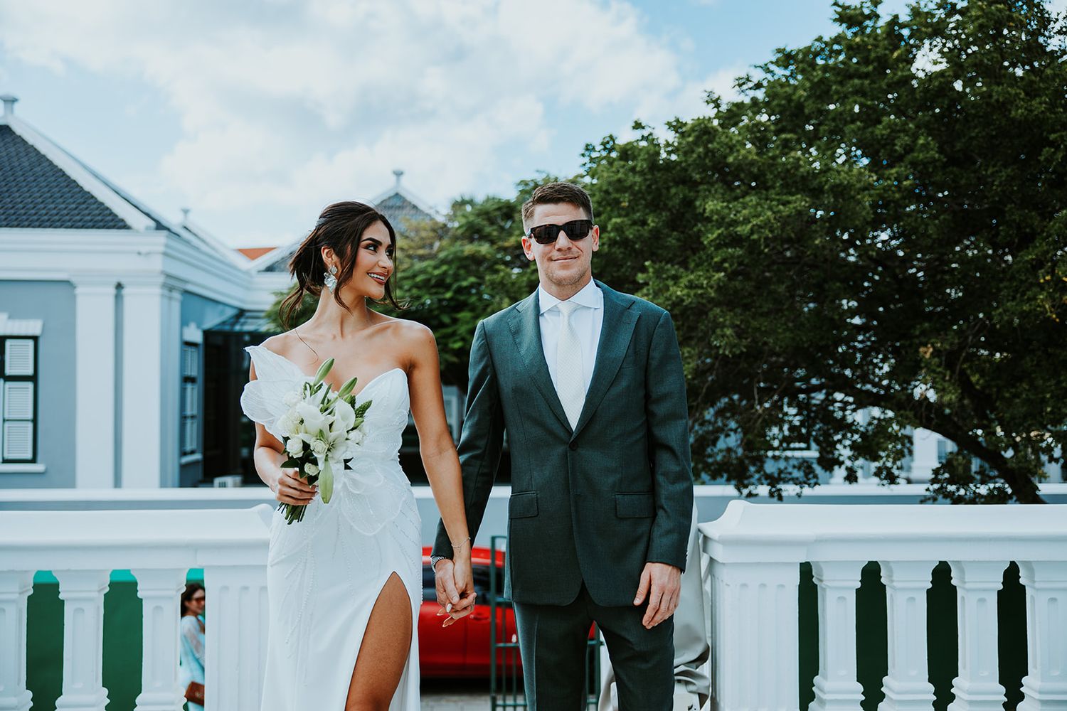 A couple poses on a white porch railing surrounded by greenery during a wedding photoshoot.