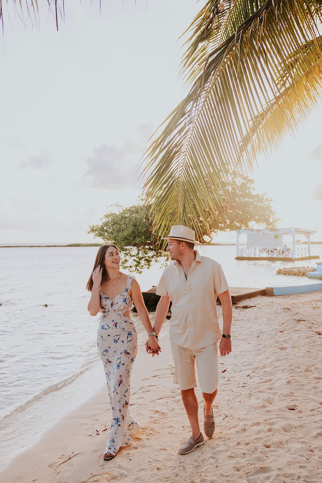 A couple holding hands walks along a tropical beach at sunset with palm trees and ocean views in the background.