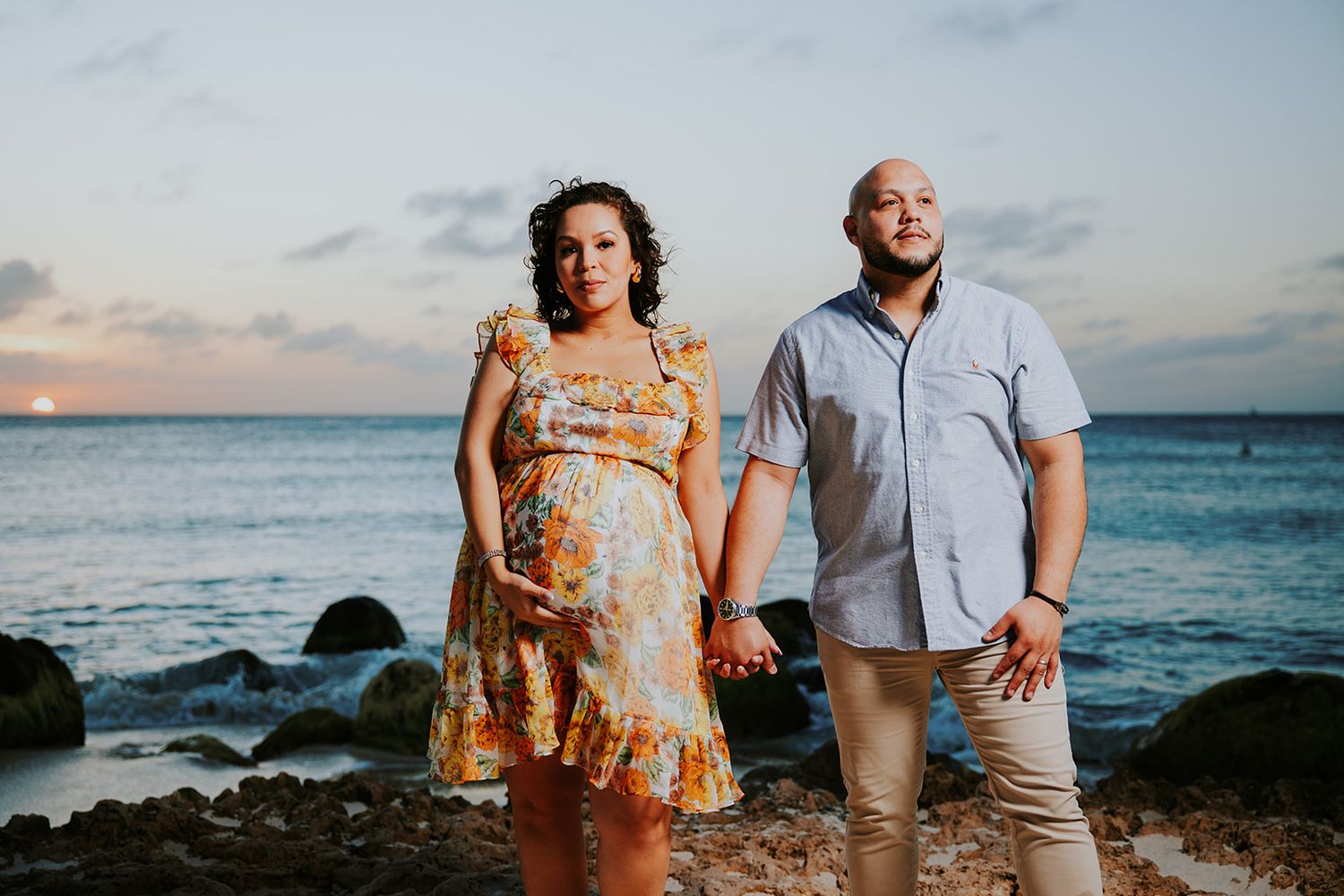 A couple stands together at a rocky beach during sunset while waves crash against the Hawaiian coastline.