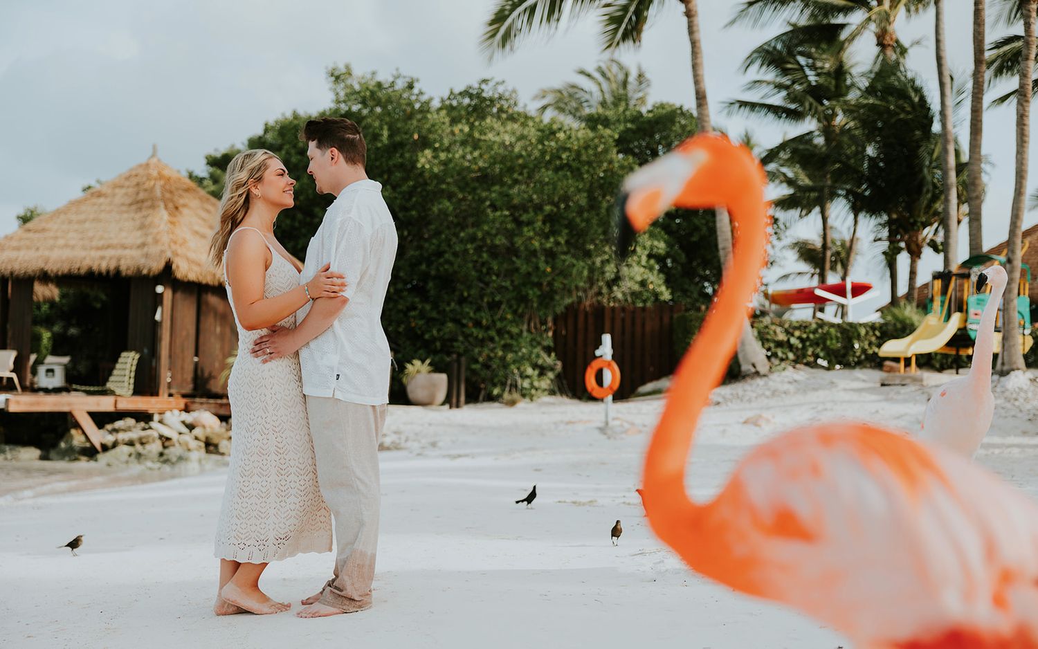 A couple poses together on a tropical beach with pink flamingo decorations at sunset.