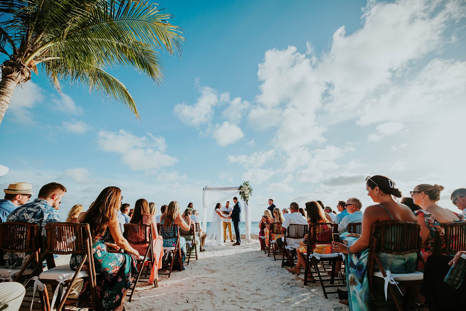 A beach wedding ceremony features guests seated on white chairs beneath palm trees and a blue Caribbean sky.