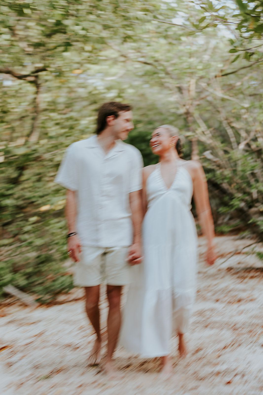 Couple in light summer clothing walk hand in hand through sunlit wooded path.