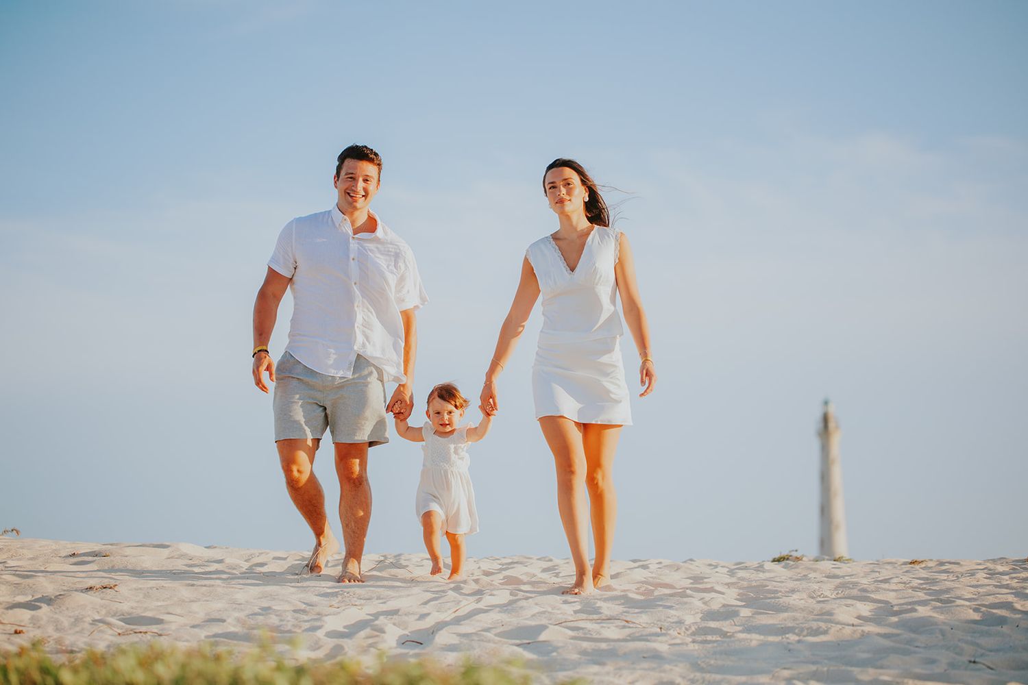 Happy family with toddler walking on sandy beach in white outfits, lighthouse in background on sunny day.