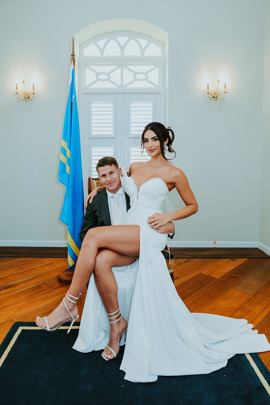 A bride in a white wedding dress sits on a dark blue carpet while adjusting her heels in an elegant room.