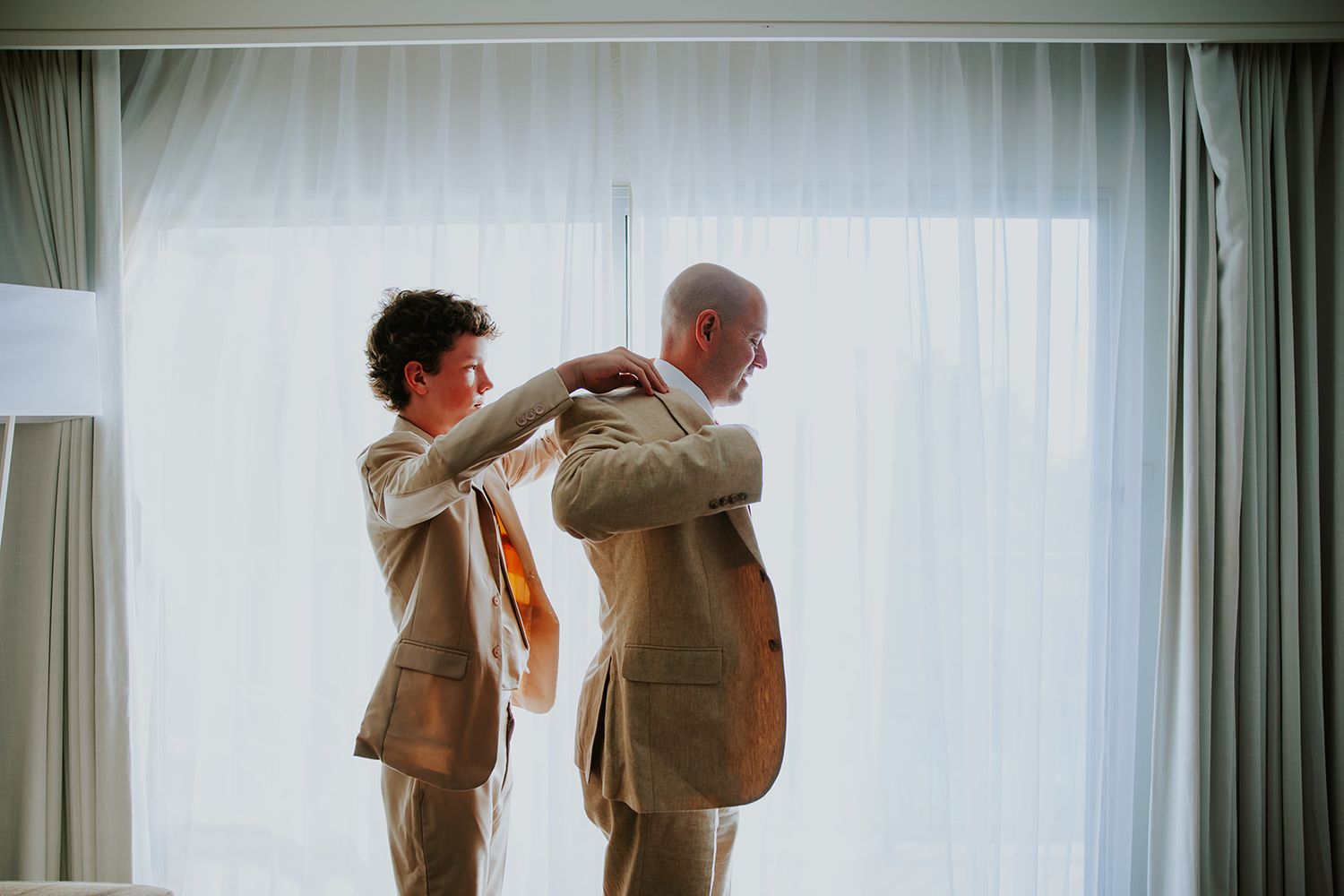 Two people in matching beige suits help each other get dressed in front of a window with sheer curtains.
