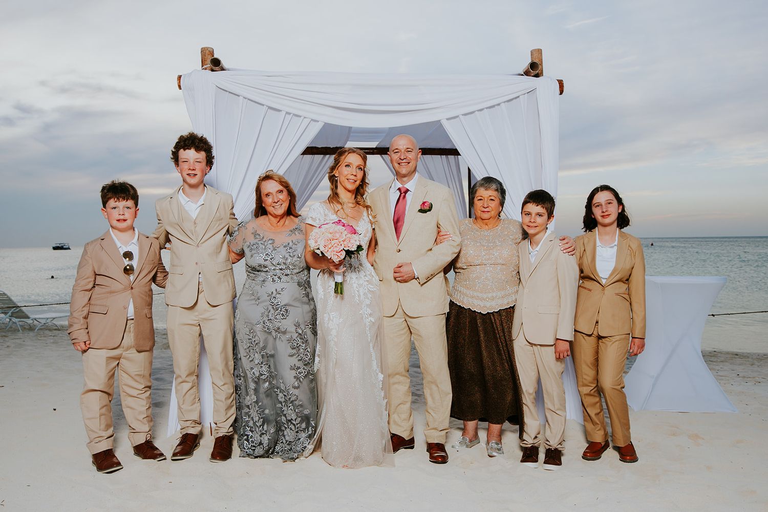 Wedding party in neutral colored attire poses together on a scenic beach at sunset.