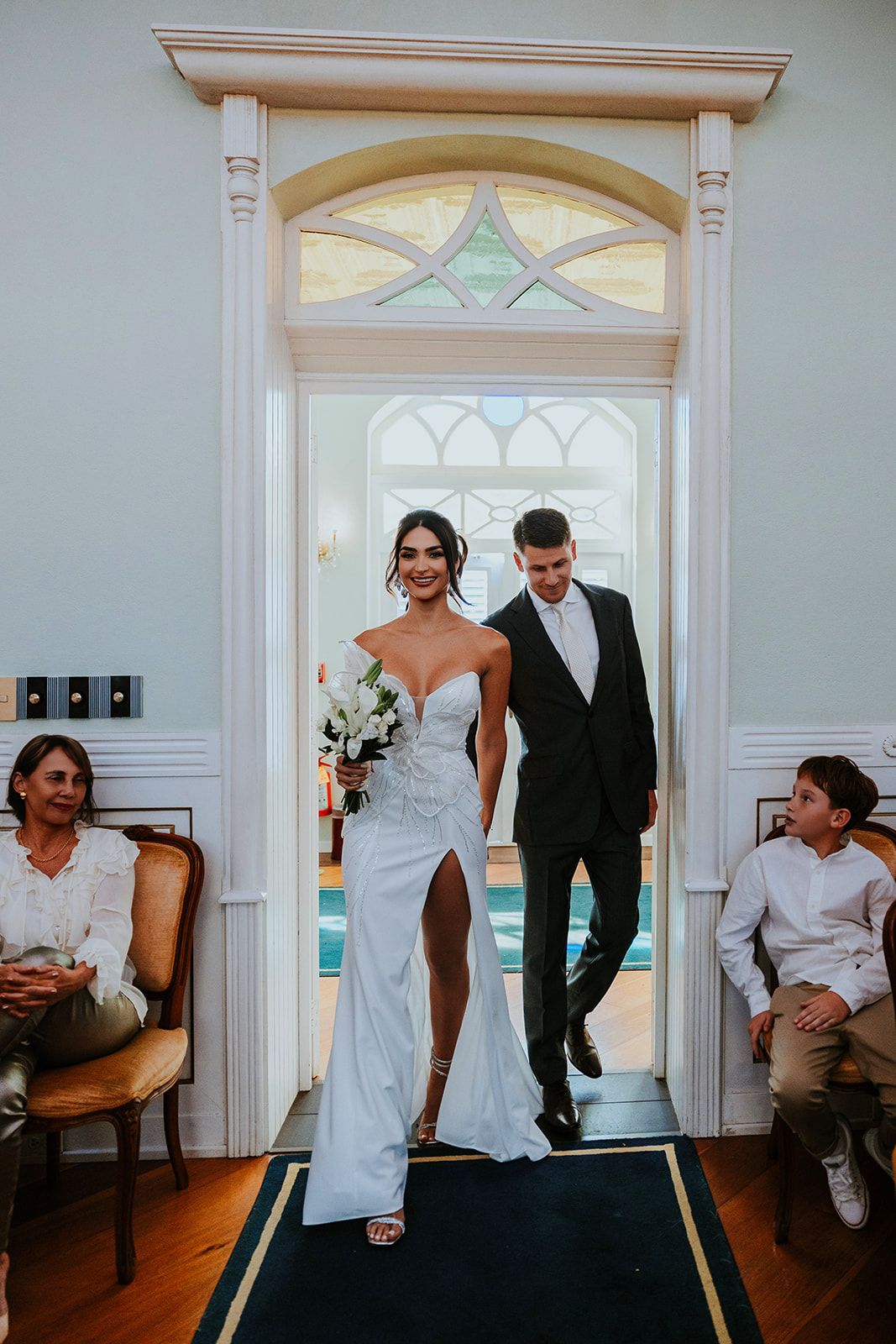 A wedding couple poses for artistic portraits in a bright white hallway with elegant arched windows and doorways.