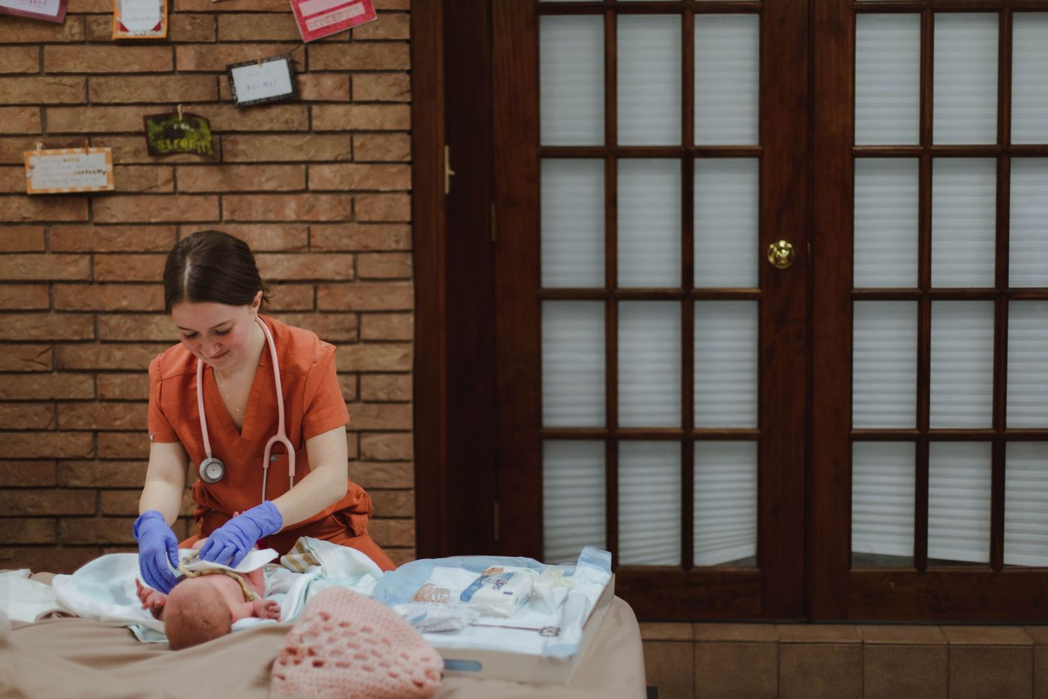 A nurse in orange scrubs examines a newborn baby on a changing table in a warmly decorated home setting.