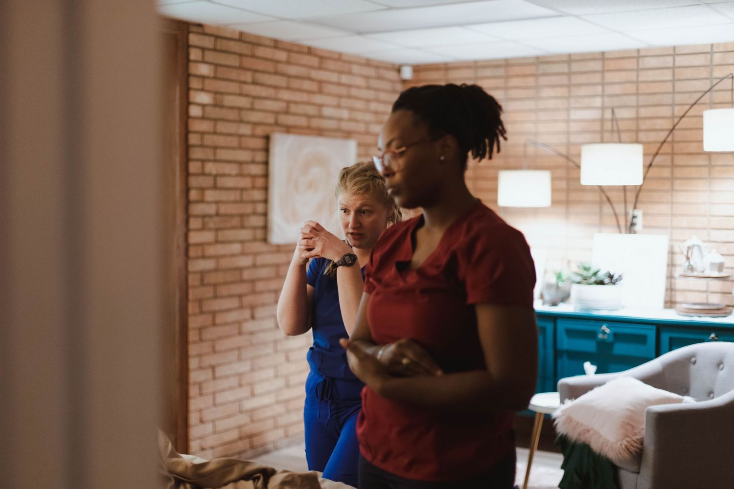 Midwife in blue scrubs consulting with new mother holding newborn baby in cozy home setting.