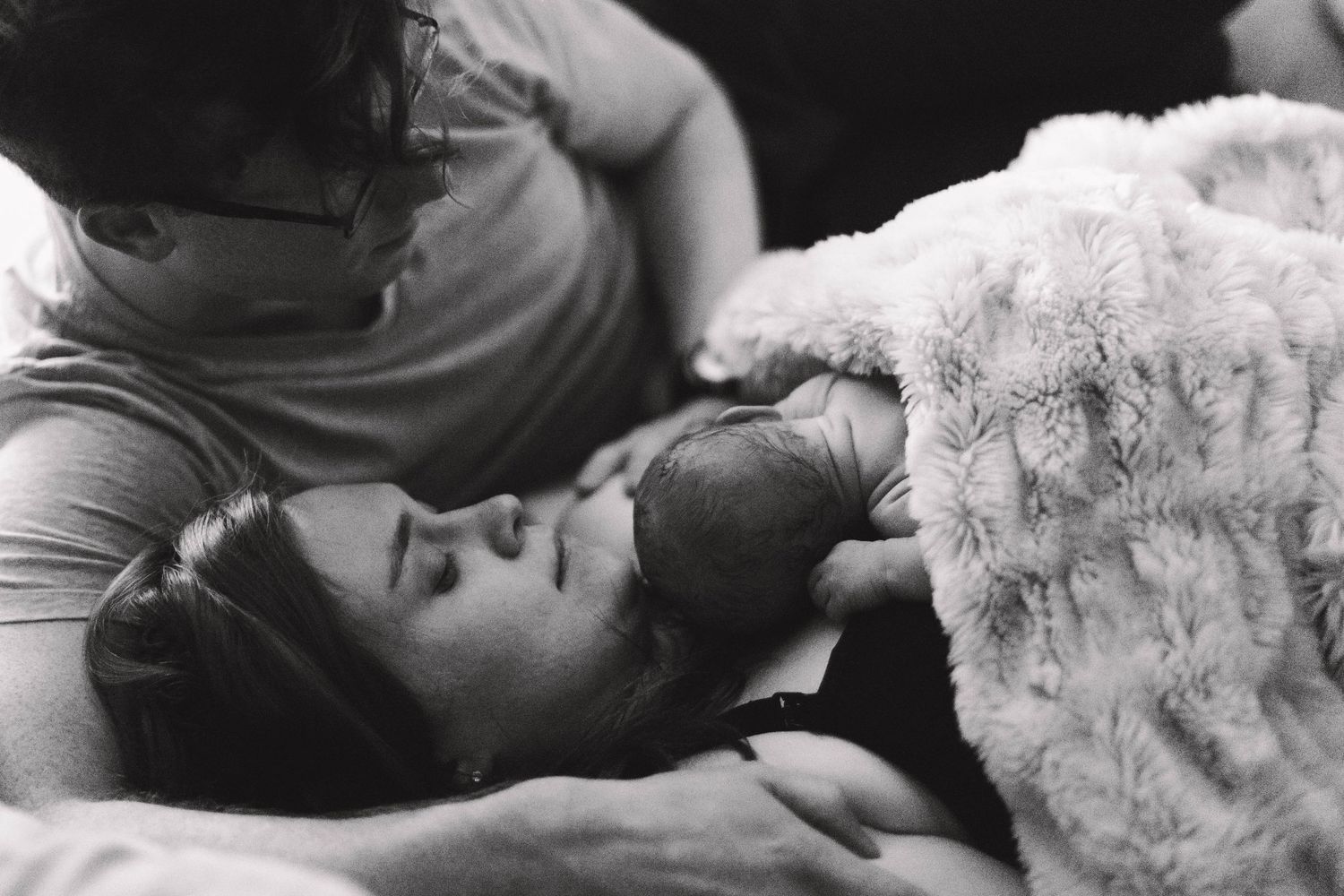 Black and white photo of parents sharing a tender moment together with their newborn baby in bed.